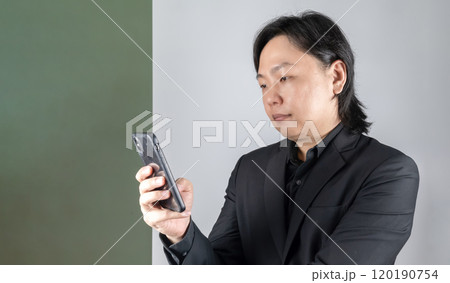 The portrait of a young Asian man dressed in a sleek black suit, engrossed in his mobile device 120190754