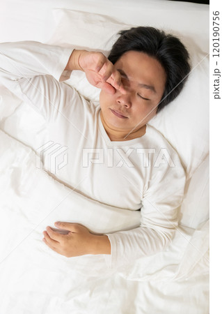 A drowsy young man in white nightwear rubs his eye while laying down on a bed in a house. A man tucks away in a soft white blanket and put a hand on his stomach. A drowsy young man in white nightwear rubs his eye while laying down on a bed in a house. A man tucks away in a soft white blanket and put a hand on his stomach. 120190756