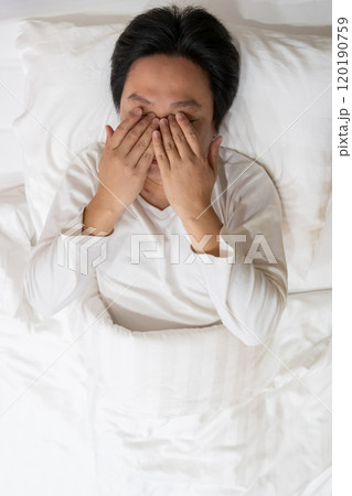 A drowsy young man in white nightwear rubs his eye while laying down on a bed in a house. A man tucks away in a soft white blanket and put a hand on his stomach. A drowsy young man in white nightwear rubs his eye while laying down on a bed in a house. A man tucks away in a soft white blanket and put a hand on his stomach. 120190759