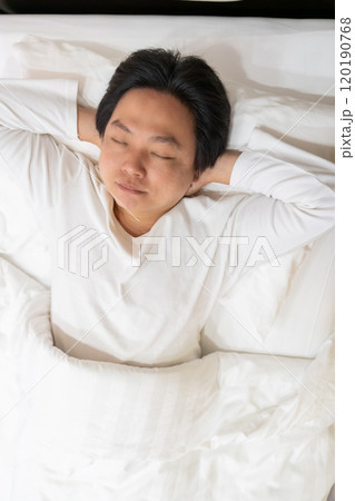 Enjoying a restful sleep, an Asian man finds comfort in his white long-sleeved cotton pajama shirt as he lies on a soft bed, nestled under a gentle blanket and supported by a plush pillow. 120190768