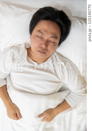 Enjoying a restful sleep, an Asian man finds comfort in his white long-sleeved cotton pajama shirt as he lies on a soft bed, nestled under a gentle blanket and supported by a plush pillow. Enjoying a restful sleep, an Asian man finds comfort in his white long-sleeved cotton pajama shirt as he lies on a soft bed, nestled under a gentle blanket and supported by a plush pillow. 120190770