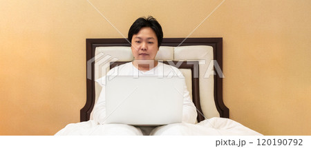 Work from home, a freelancer man leaning against the bed and using his laptop to work online on a bed. Work from home, a freelancer man leaning against the bed and using his laptop to work online on a bed. 120190792