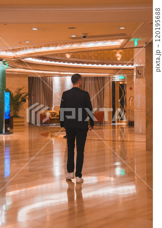 A man in a dark suit walks through a luxurious lobby with marble floors, modern decor, and intricate lighting fixtures in Shanghai. 120195688