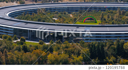 The image captures an aerial view of Apple Park in Cupertino, California, featuring the circular building, lush greenery, and a colorful rainbow arch. 120195788