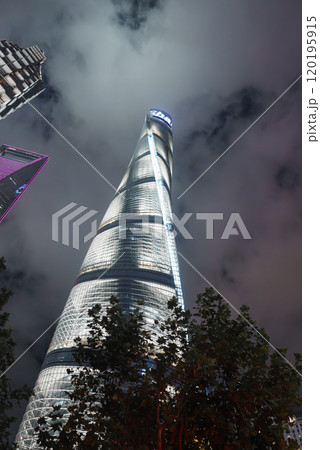 Shanghai Tower stands illuminated against a cloudy night sky, with its spiraling design highlighted. Silhouettes of trees and another skyscraper are visible. 120195915