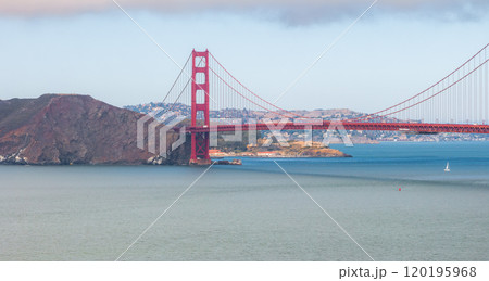 The Golden Gate Bridge spans the San Francisco Bay with its International Orange color. A sailboat navigates the blue waters, with Marin County hills visible. 120195968