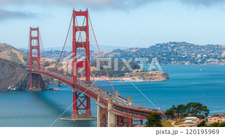 The Golden Gate Bridge spans the blue waters of San Francisco Bay, with its International Orange color contrasting against rolling hills and a cloudy sky. 120195969