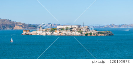 Alcatraz Island with its historic penitentiary and watchtower is surrounded by San Francisco Bay's blue waters. A sailboat and Marin Headlands are visible. 120195978