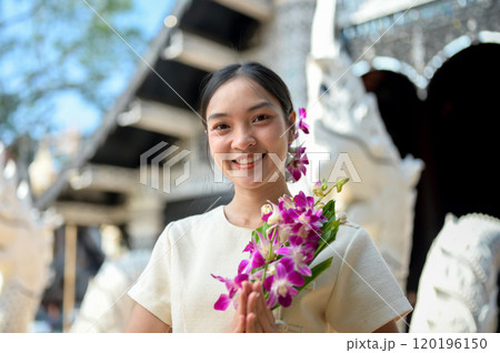A beautiful, smiling Asian women stand in a temple, holding flowers in a prayer posture. 120196150