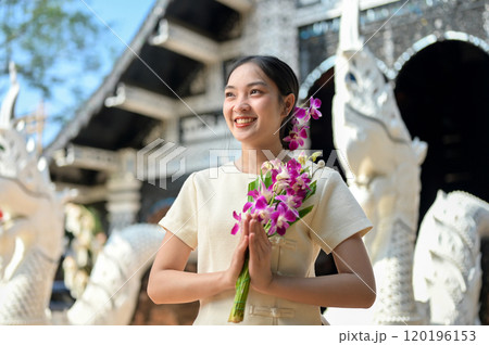 A smiling Asian women in traditional dresses stand in a temple, holding flowers in a prayer posture. 120196153
