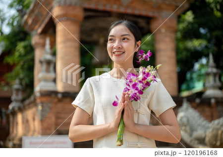 A charming Asian woman stands outdoors in a temple, holding a flower in a praying posture. 120196168