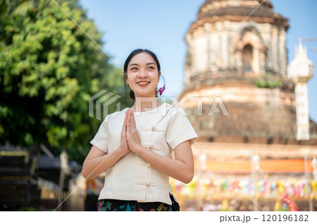 A charming Asian woman in a traditional Thai Lanna dress stands outdoors in a praying posture. 120196182