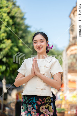 A charming Asian woman in a traditional Thai Lanna dress stands outdoors in a praying posture. 120196183