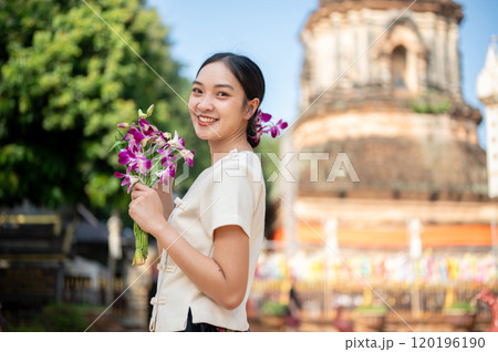 A charming Asian woman in a traditional dress smiling at the camera with a pagoda in the background. 120196190
