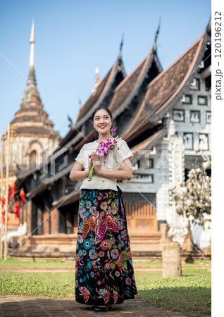 Charming Asian woman in a traditional dress stands outdoors with an ancient temple in the background 120196212