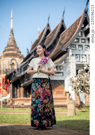 Charming Asian woman in a traditional dress stands outdoors with an ancient temple in the background 120196213