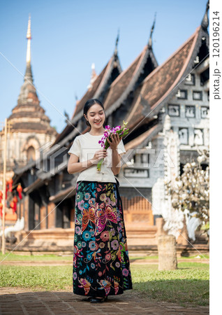 A charming Asian woman with a temple in the background, admiring a flower bouquet in her hand. 120196214