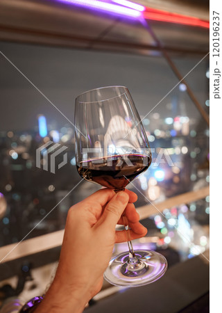 A hand holds a glass of red wine with Shanghai's vibrant cityscape in the background. Blurred colorful lights highlight the city's modern architecture. 120196237
