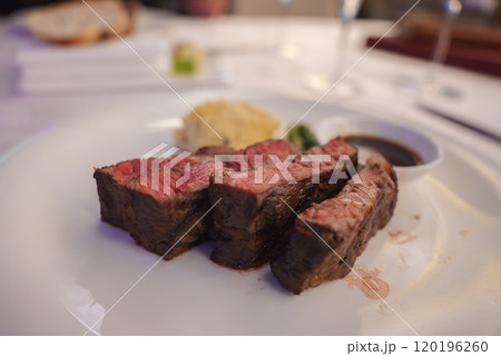 A plated dish features three slices of medium rare steak with a seared exterior, accompanied by mashed potatoes, green garnish, and a side of sauce. A plated dish features three slices of medium rare steak with a seared exterior, accompanied by mashed potatoes, green garnish, and a side of sauce. 120196260