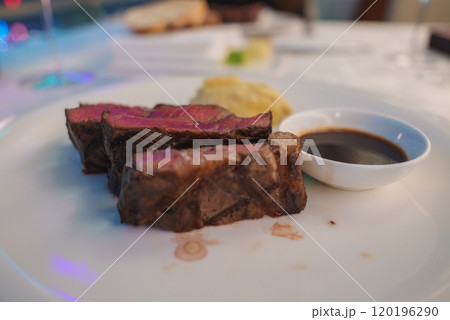 A plate displays slices of medium rare steak with a pink interior and seared exterior, accompanied by a small dish of dark sauce. Background is softly blurred. A plate displays slices of medium rare steak with a pink interior and seared exterior, accompanied by a small dish of dark sauce. Background is softly blurred. 120196290