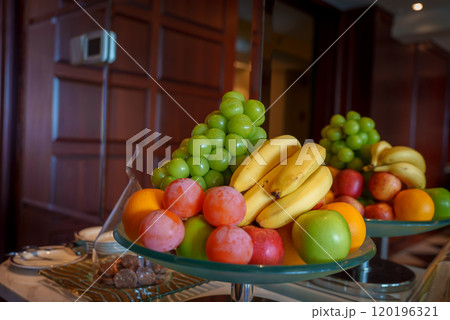 A colorful arrangement of bananas, grapes, apples, oranges, and plums on a glass platter indoors, set against a wooden paneled wall. 120196321