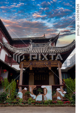 A traditional Chinese building in Shanghai features an ornate, curved roof with intricate carvings. Lush greenery and vibrant flowers adorn the foreground. A traditional Chinese building in Shanghai features an ornate, curved roof with intricate carvings. Lush greenery and vibrant flowers adorn the foreground. 120196326