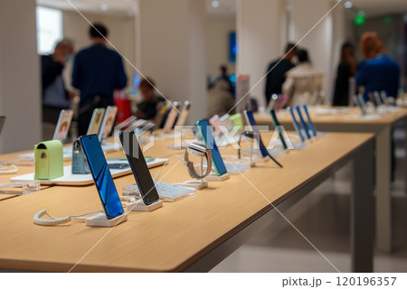 A variety of smartphones are displayed on a sleek wooden table in a well lit, minimalist electronics store. People are visible in the background. A variety of smartphones are displayed on a sleek wooden table in a well lit, minimalist electronics store. People are visible in the background. 120196357