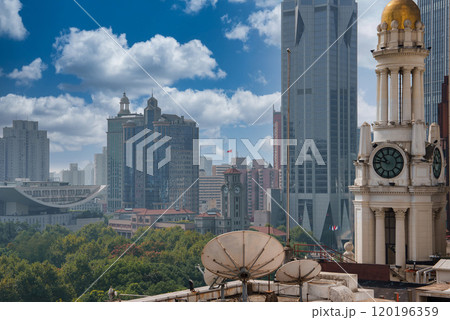 View of Shanghai's skyline featuring a clock tower with a golden dome, modern skyscrapers, lush greenery, and rooftop satellite dishes under a blue sky. 120196359