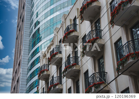 A sleek skyscraper with reflective glass stands beside a classic building with ornate balconies and red flowers, set against a clear blue sky in Shanghai. A sleek skyscraper with reflective glass stands beside a classic building with ornate balconies and red flowers, set against a clear blue sky in Shanghai. 120196369