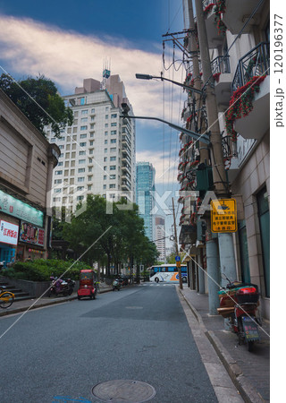 A quiet street in Shanghai, China, features buildings with flower adorned balconies, parked scooters, and a red tuk tuk, set against modern skyscrapers. 120196377