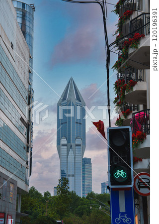 Tomorrow Square's angular design stands out against a cloudy sky, framed by a traffic light with a bicycle signal and a building with red flowers. 120196391