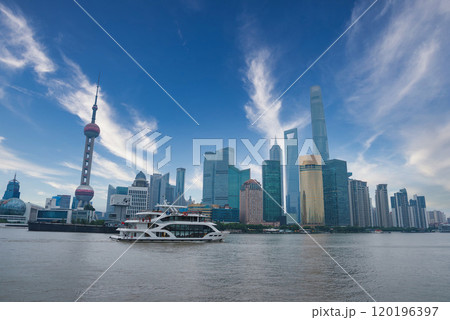 The image captures Shanghai's skyline, featuring the Bund, Oriental Pearl Tower, and Shanghai Tower. A boat cruises on the Huangpu River under a clear sky. 120196397