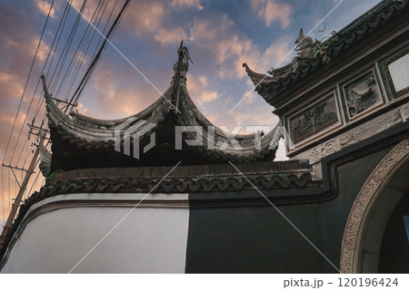 Traditional Chinese building with curved roofs and carvings set against a sunset sky. Power lines add a modern contrast in Shanghai's old town. 120196424