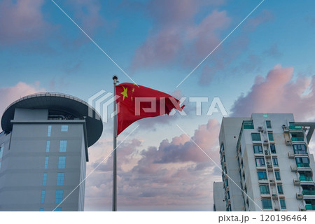 A Chinese flag waves prominently with a pink and blue sky backdrop. A modern building with a circular rooftop and a high rise residential building are visible. 120196464