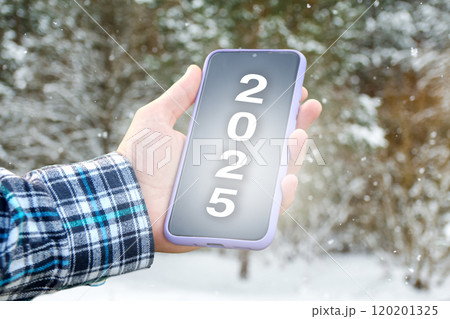 a hand holding a smartphone displaying the number 2025 on the screen, symbolizing the New Year. The background features a winter scene 120201325