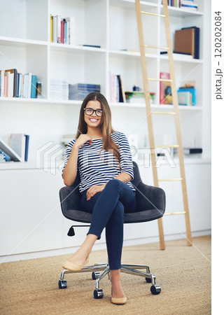 She got such a winning smile. Portrait of a smiling young woman wearing glasses sitting in front of bookshelves at home. 120202084