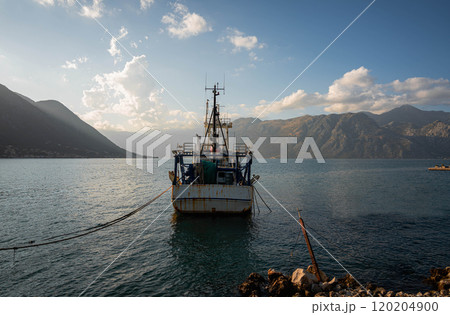 A weathered fishing trawler sits anchored near the shore, its rusted hull contrasting against the serene coastal landscape with towering mountains and a peaceful sky at dusk. A weathered fishing trawler sits anchored near the shore, its rusted hull contrasting against the serene coastal landscape with towering mountains and a peaceful sky at dusk. 120204900