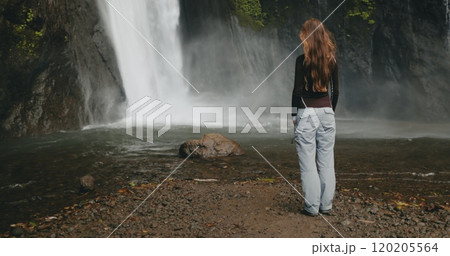 Tourist woman looking powerful Bali waterfall, water cascading down rocks. Long hair blows in wind. Tourism, trekking, hiking, relax, active lifestyle, summer holiday vacation. Slow back view Tourist woman looking powerful Bali waterfall, water cascading down rocks. Long hair blows in wind. Tourism, trekking, hiking, relax, active lifestyle, summer holiday vacation. Slow back view 120205564
