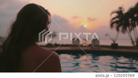Woman relaxes near swimming pool at sunset. Long hair brunette girl back view enjoys colorful sky reflected in water, palm trees in background. Tropical resort, travel, exotic vacation in Thailand Woman relaxes near swimming pool at sunset. Long hair brunette girl back view enjoys colorful sky reflected in water, palm trees in background. Tropical resort, travel, exotic vacation in Thailand 120205569
