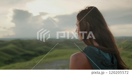 Young woman stands on a hill in indonesia, watching the sunset over lush green hills, painting the sky with orange and pink hues Young woman stands on a hill in indonesia, watching the sunset over lush green hills, painting the sky with orange and pink hues 120205572