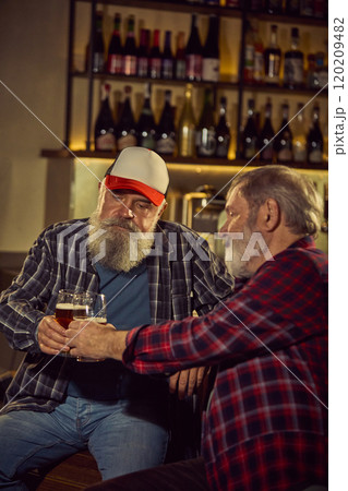 Two elderly men in checkered shirts sitting at modern cozy bar, drinking beer, involved in conversation, and enjoying sport translation 120209482