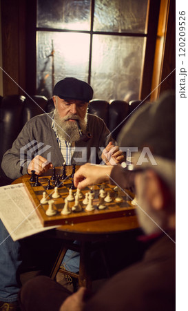 Bearded elderly man in a cap, smoking tobacco pipe and playing chess with his friend in cozy vintage bar Bearded elderly man in a cap, smoking tobacco pipe and playing chess with his friend in cozy vintage bar 120209526