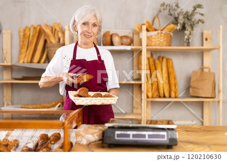 Friendly female baker in apron putting hot delicious croissants to showcase for sale Friendly female baker in apron putting hot delicious croissants to showcase for sale 120210630
