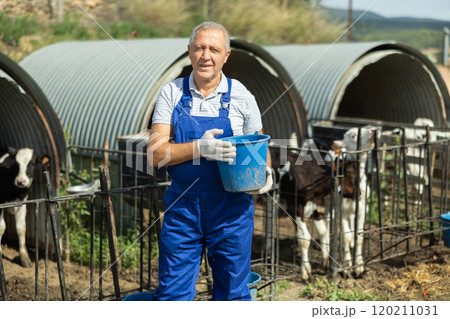 Senior male employee stands near street cow pen with bucket 120211031