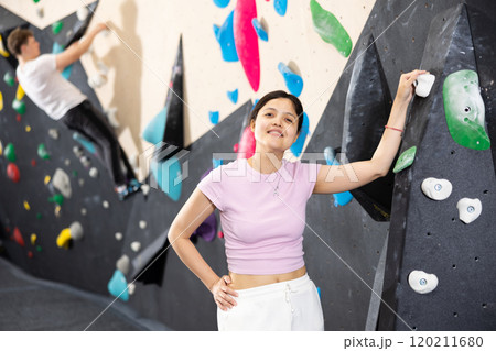 Young smiling Asian girl coach stands at climbing wall and leans holding on to hook. 120211680