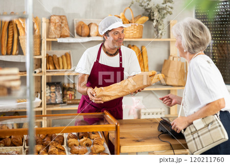 Man entrepreneur, owner of baker puts baguettes in paper bag, serves senior female client Man entrepreneur, owner of baker puts baguettes in paper bag, serves senior female client 120211760