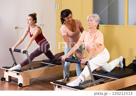 Two active women perform an exercise using a reformer bed Two active women perform an exercise using a reformer bed 120212513