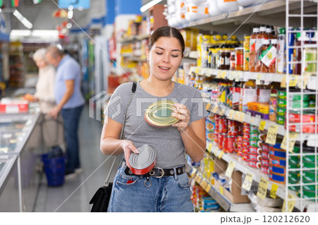 Young woman purchaser choosing tinned fish in big supermarket Young woman purchaser choosing tinned fish in big supermarket 120212620