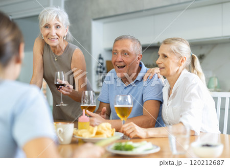 Cheerful elderly women and man gathering around kitchen table Cheerful elderly women and man gathering around kitchen table 120212687