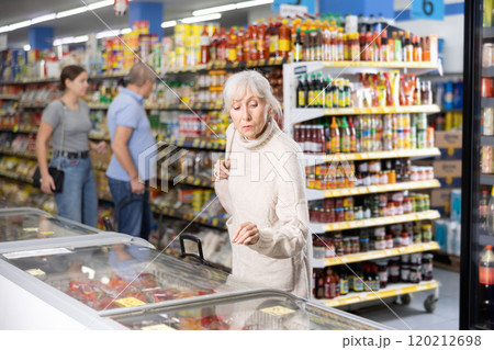 Old woman purchaser choosing frozen product in big supermarket 120212698